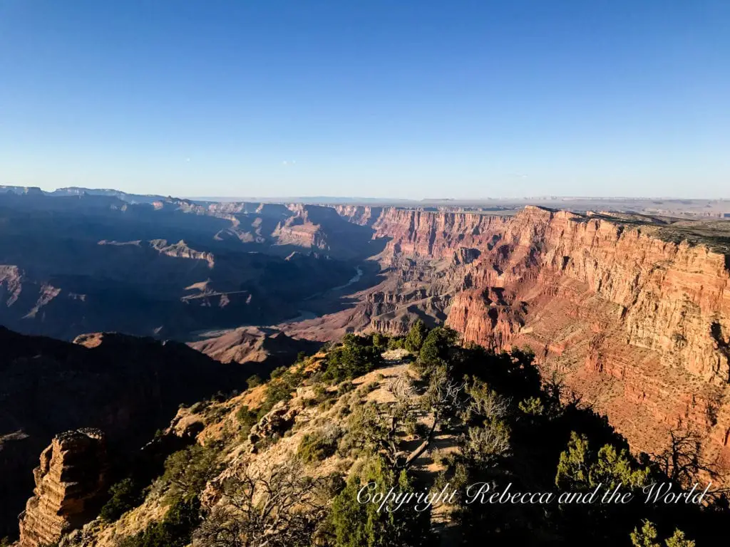 Expansive view of the Grand Canyon with a clear blue sky overhead. Jagged cliffs and deep valleys define the landscape, with layers of red, brown, and orange rock. The Colorado River meanders in the distance.