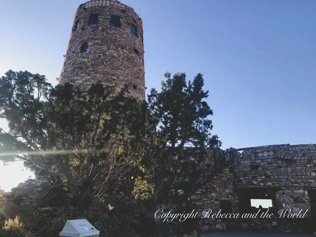 Stone watchtower with a conical roof, situated at the Grand Canyon's edge. The structure is made of natural rock, blending with the environment. Trees partially obscure the view, with sunlight filtering through.