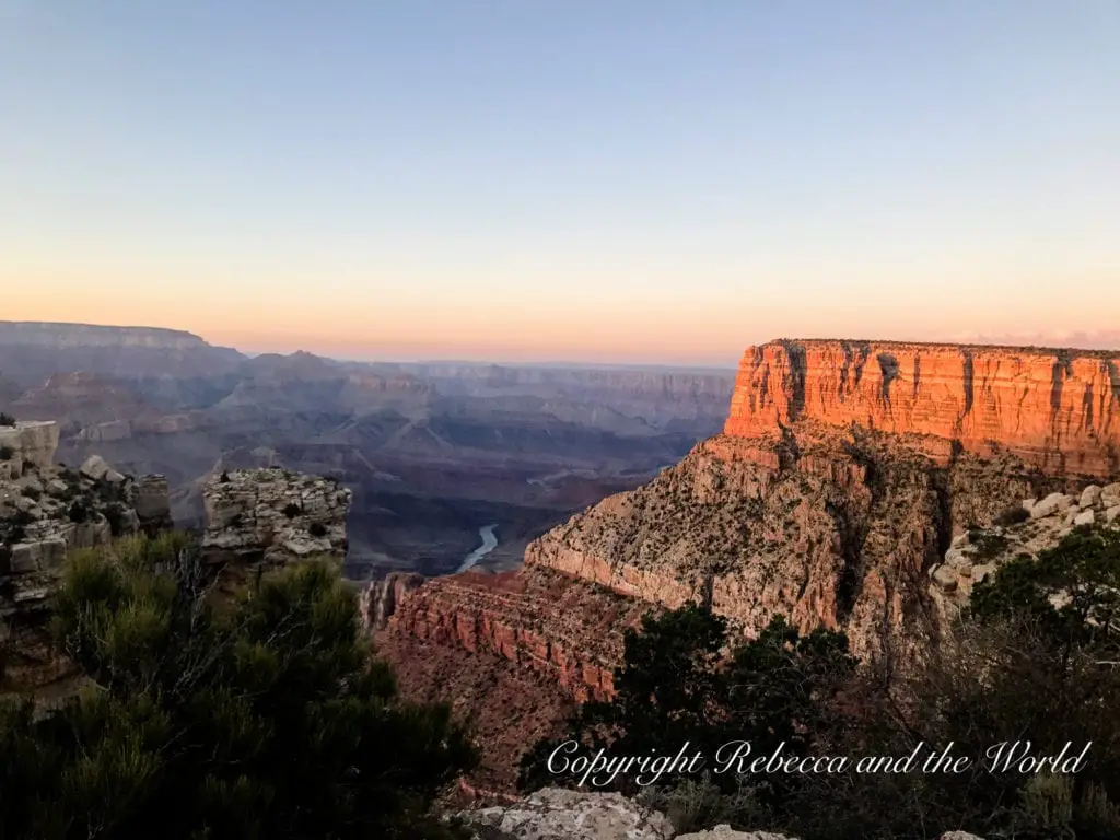 Twilight at the Grand Canyon, with the horizon displaying soft pastel colors. The canyon walls glow with a warm orange hue, highlighting their textured surfaces. The river below is faintly visible.