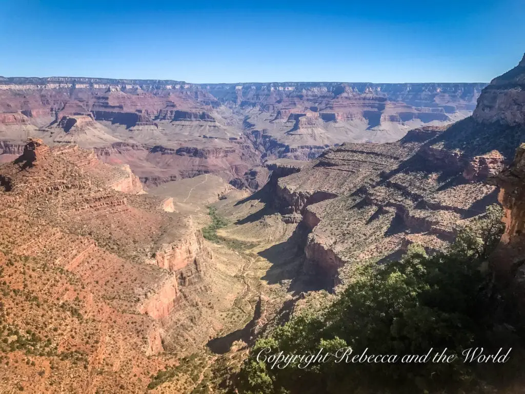 Bright daylight view over the Grand Canyon. Sharp shadows accentuate the depth and contours of the terrain. Rich reds and browns dominate the scene, with sparse green vegetation throughout.