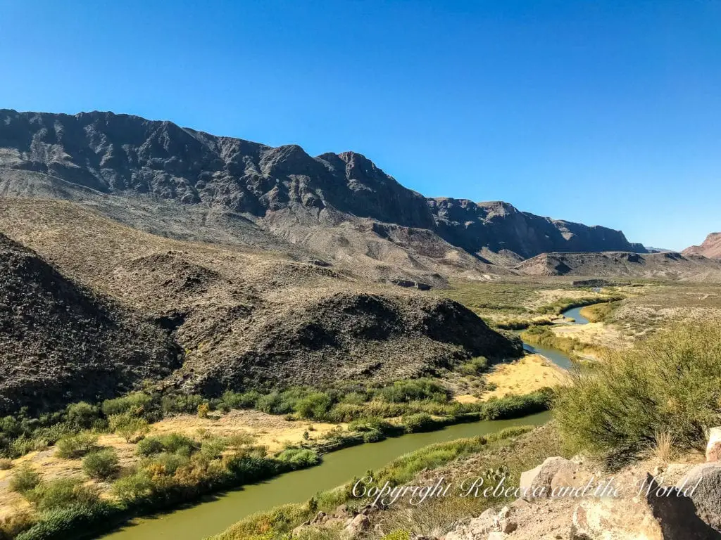 A serene view of a meandering river cutting through a desert landscape with rugged mountains in the background under a clear blue sky. The Rio Grande separates Texas from Mexico and the River Road Drive is a must-do in West Texas.