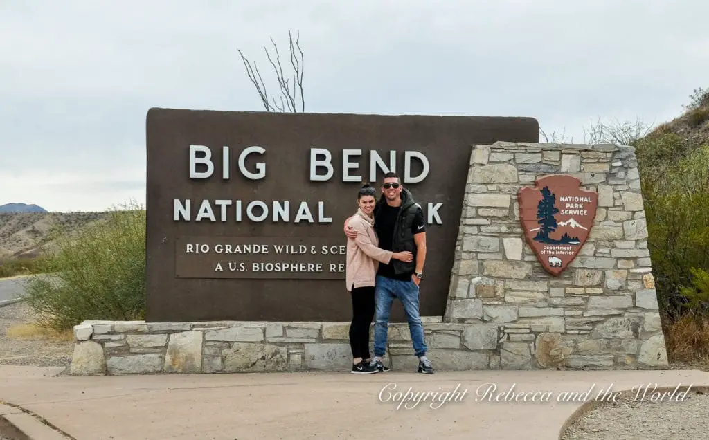 A couple - the author and her husband - poses in front of the "Big Bend National Park" entrance sign, featuring bold lettering and the National Park Service emblem on a stone plinth.