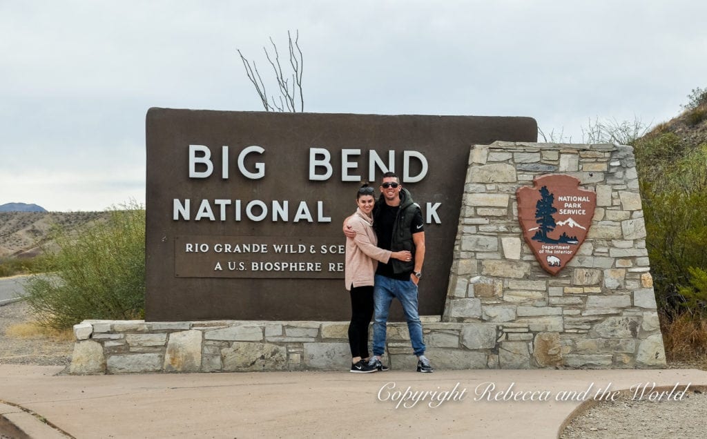 A couple - the author and her husband - poses in front of the "Big Bend National Park" entrance sign, featuring bold lettering and the National Park Service emblem on a stone plinth.