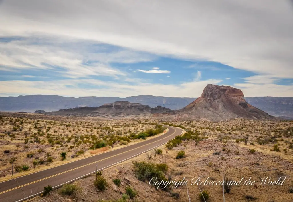 A winding road traverses a desolate landscape with sparse vegetation, leading towards a distinctive flat-topped mesa under a wide sky with wispy clouds. Big Bend National Park is a must-visit on a West Texas road trip.