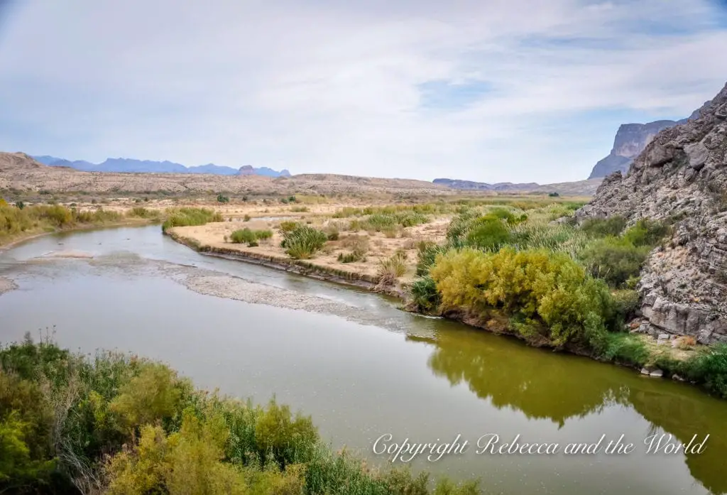 A panoramic view of a winding river through a desert landscape, with lush greenery along the banks contrasting with the surrounding arid terrain. This is the Rio Grande running through Big Bend National Park in West Texas.
