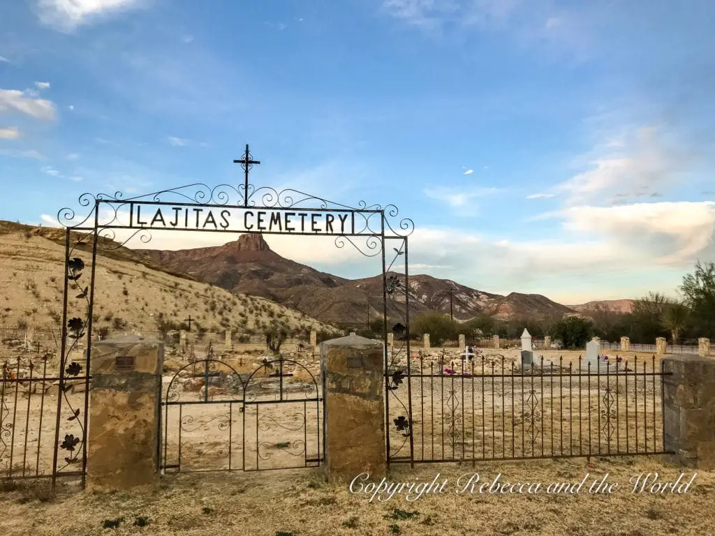 The ornate metal gate of the "Lajitas Cemetery" set against a dusky sky, with arid hills in the background and various grave markers visible beyond the gate.
