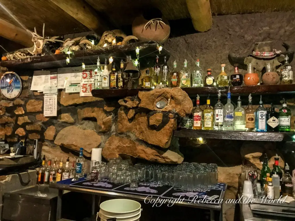 An intimate and rustic bar setting inside a stone structure, adorned with eclectic decorations and a variety of liquor bottles on wooden shelves. This is La Kiva, a restaurant and bar inside a cave just outside of Terlingua in West Texas.
