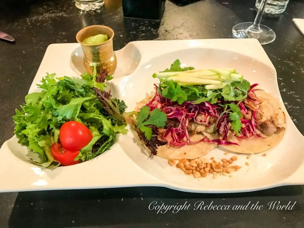 A plate with a fresh taco topped with red cabbage and avocado, beside a side salad and a small cup of green sauce, exemplifying the local gourmet cuisine available in Fredericksburg. 