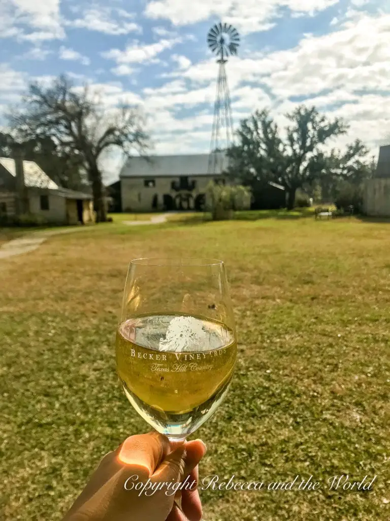 A hand holding a glass of white wine with the label 'Becker Vineyards, Texas Hill Country', with a rustic windmill and vineyard buildings blurred in the background, representing local wine tasting experiences.