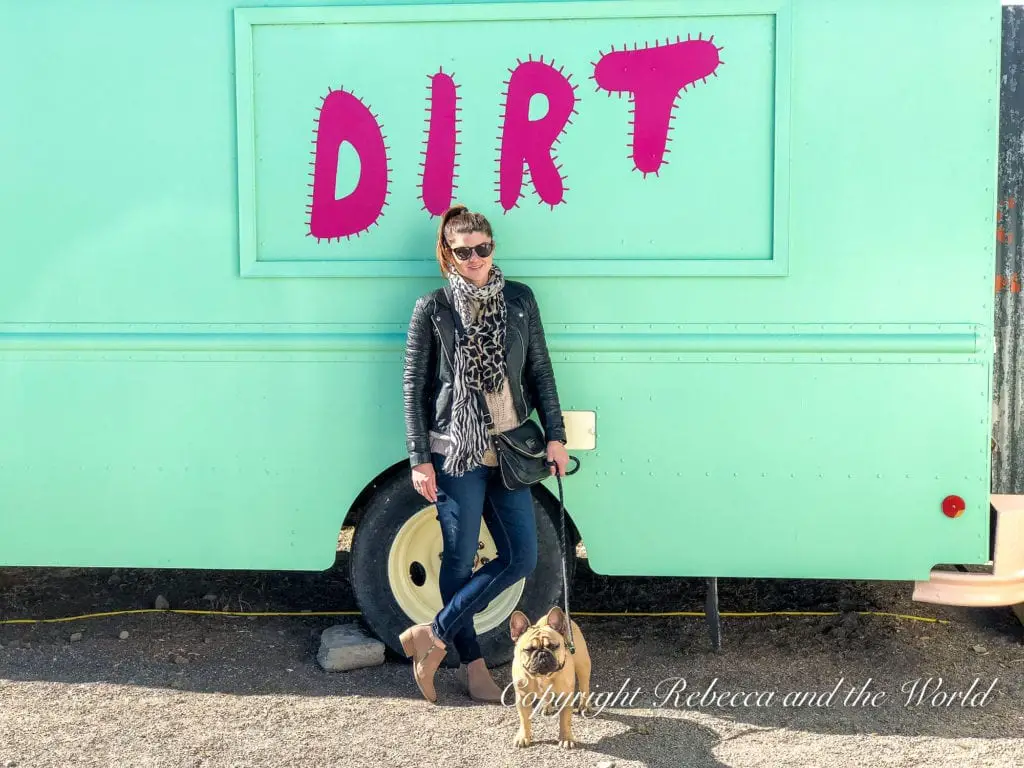 A woman - the author of this article - poses with her small French Bulldog in front of a pastel-colored trailer with the word 'DIRT' in bold pink letters, suggesting a playful side to Marfa's local scene.