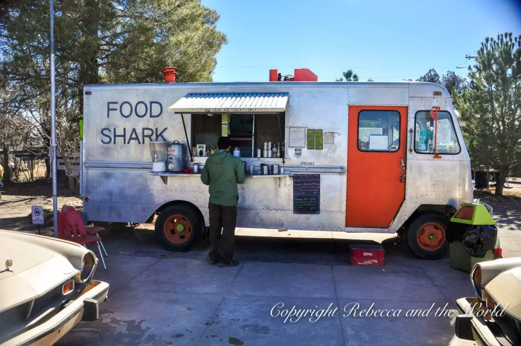 A food truck labeled 'FOOD SHARK' with a retro aesthetic is parked under trees, serving customers, with vintage cars parked nearby, suggesting a popular local eatery.