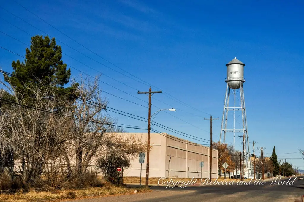 A towering white water tank labeled 'MARFA' stands prominently against a clear blue sky above a quiet street lined with power lines and a large beige building.
