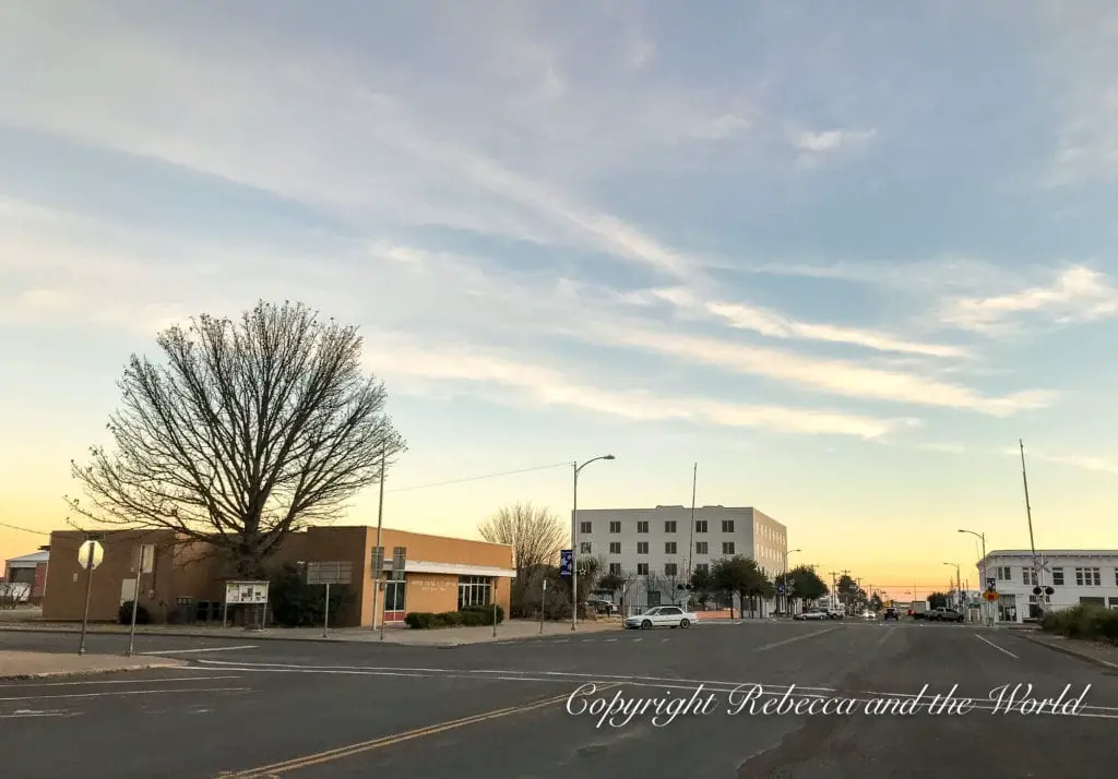 A wide street view of Marfa during twilight with soft clouds, a leafless tree silhouette, and buildings in the distance, conveying a tranquil small-town feel.