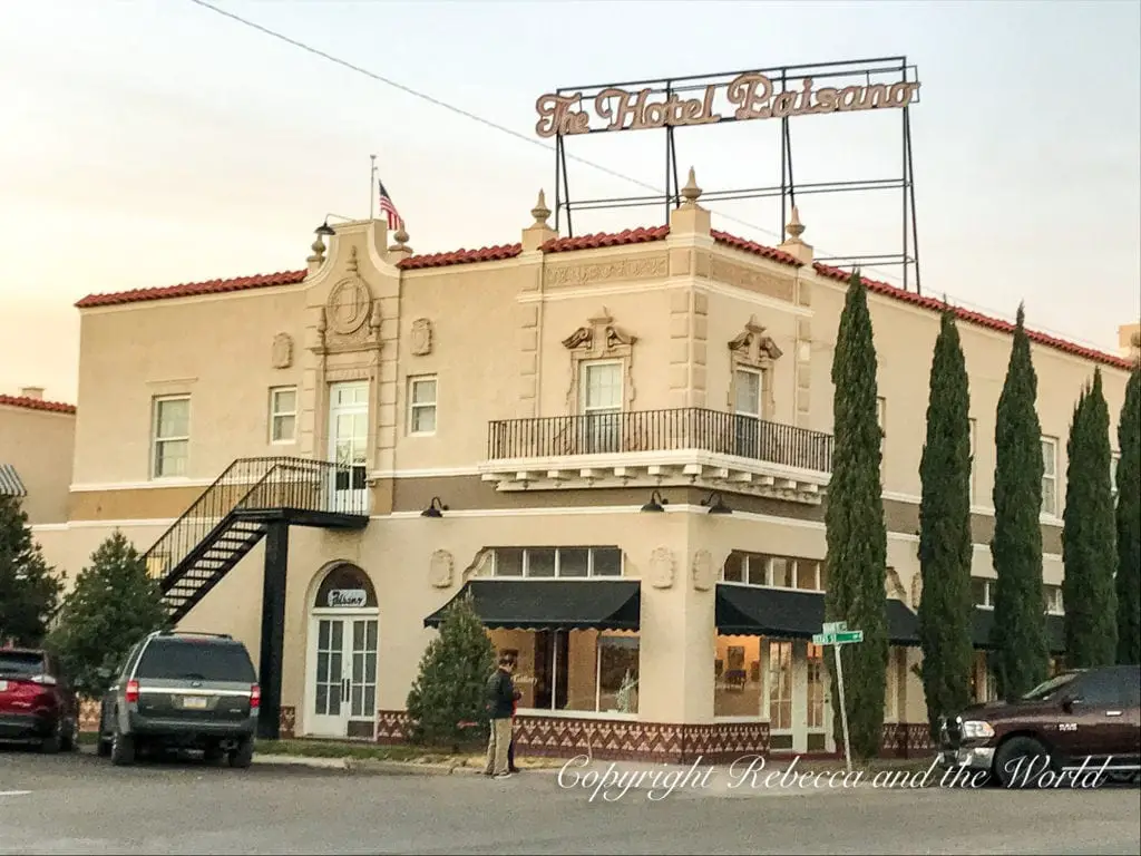 The historic Hotel Paisano with a prominent sign atop, flanked by tall cypress trees against a dusky sky, highlighting a landmark stay in Marfa. Texas.