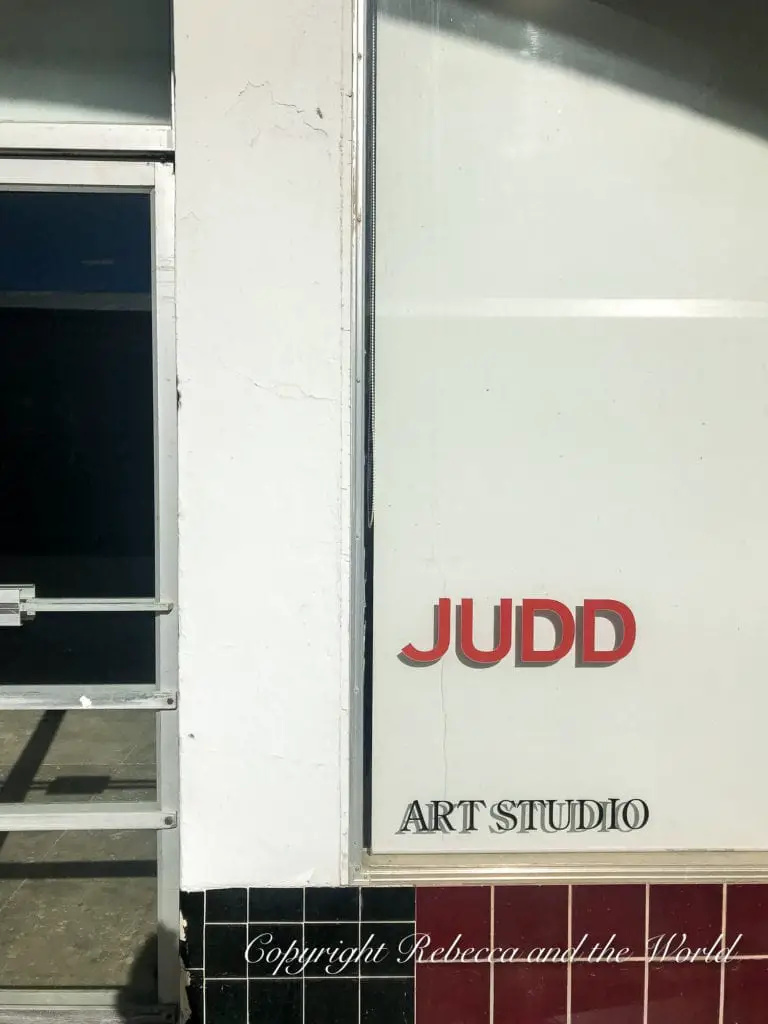 A close-up view of a corner of a white building with a door ajar, displaying the red text 'JUDD ART STUDIO' on a white wall with black and white tiles at the bottom.