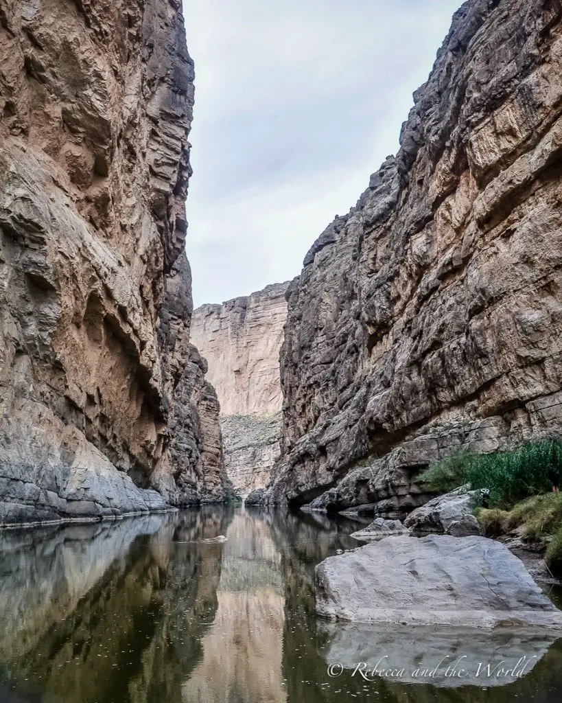 A tranquil river flowing through a narrow canyon with towering cliff walls on either side, reflecting the grey of the overcast sky. Santa Elena Canyon is an easy hike in Big Bend National Park, that leads to spectacular views.