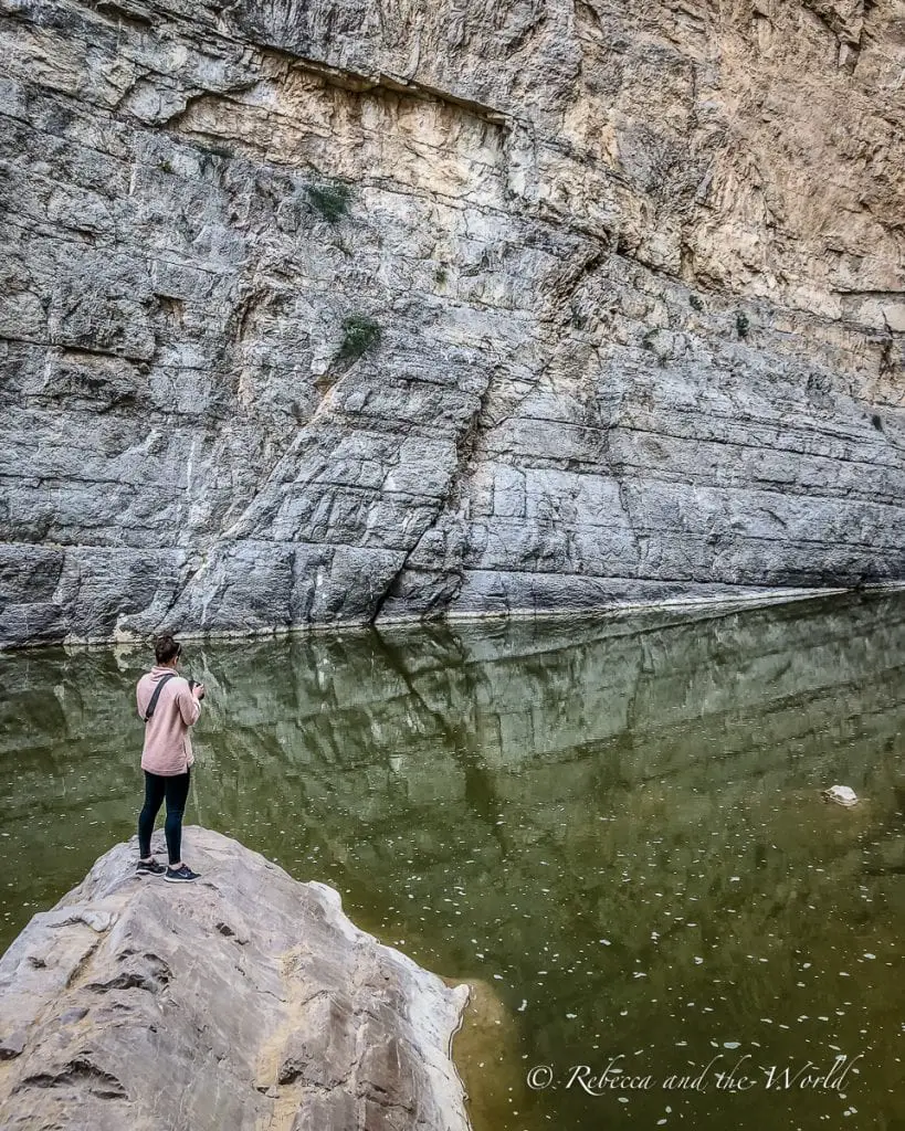 A person - the author of this article - stands on a large rock, overlooking a calm river bordered by steep canyon walls, conveying a sense of solitude and reflection. Santa Elena Canyon is an easy hike in Big Bend National Park, that leads to spectacular views.