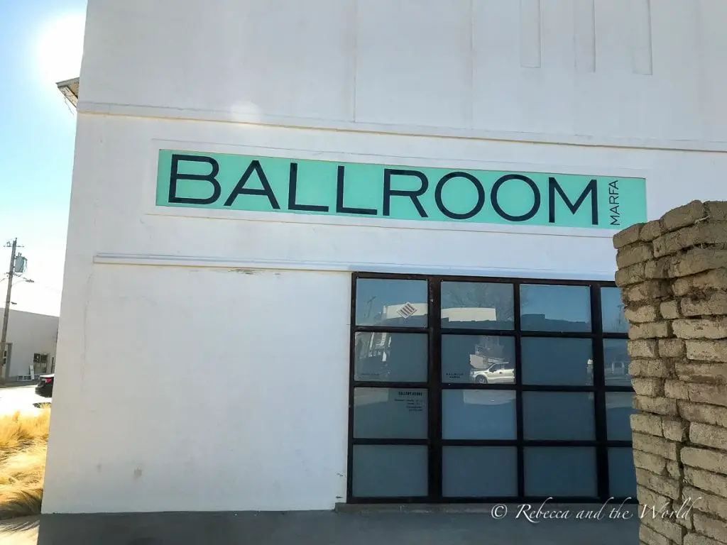 A minimalist white building with a sign that reads "BALLROOM MARFA" in mint green letters, reflecting the clear blue sky in its large windows. There are plenty of galleries to visit in Marfa to experience the towns reputation as a cultural hub.