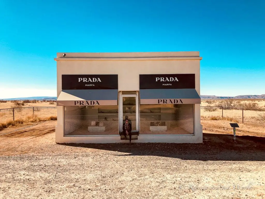 A solitary building labeled "Prada Marfa" stands alone in a barren landscape; it's a small, white, adobe-style structure with large windows showcasing luxury bags and shoes. Stopping at the Prada art installation near Marfa is one of the most popular things to do in West Texas.