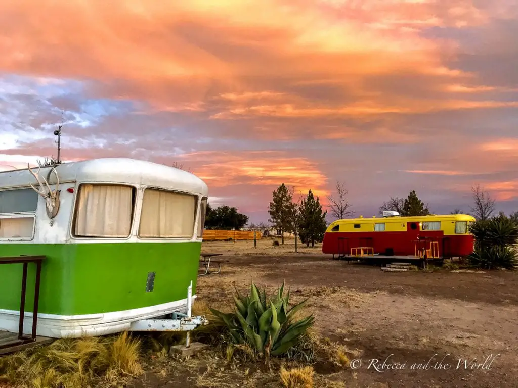 Vibrant sunset hues of pink and orange illuminate the sky over a desert scene featuring a green vintage trailer and a red retro caravan among sparse vegetation. El Cosmico is one of the best glamping spots in West Texas.