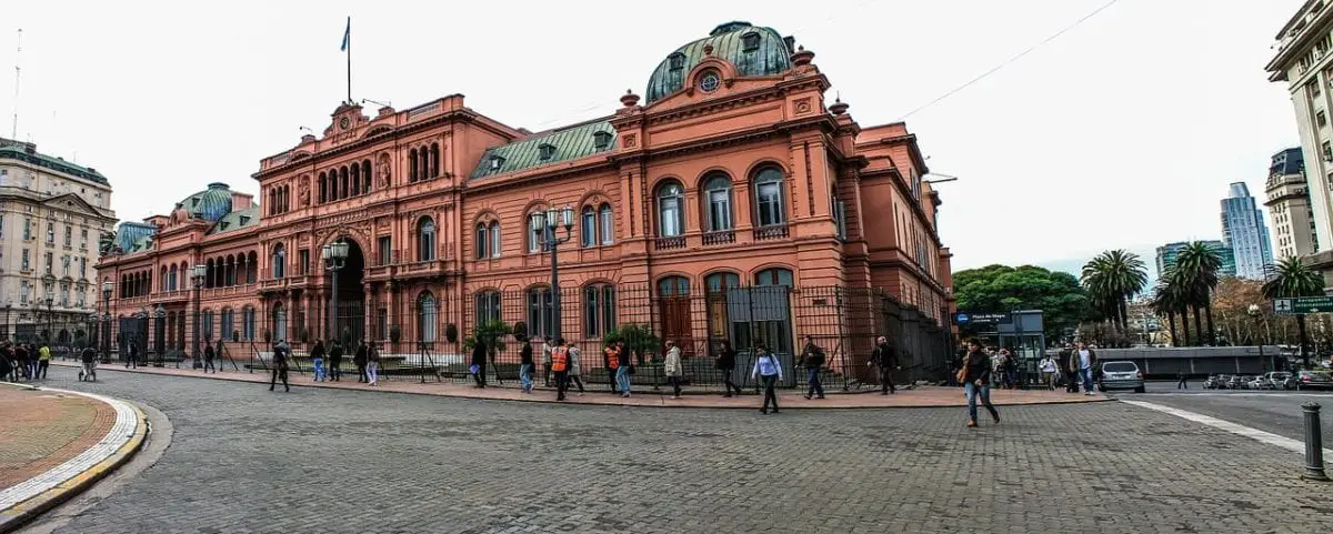 A panoramic view of La Casa Rosada, a large historic building with a pink facade, featuring multiple arches and domes. There are people walking in front, and the building is set against a backdrop of a city street with trees and overcast skies.