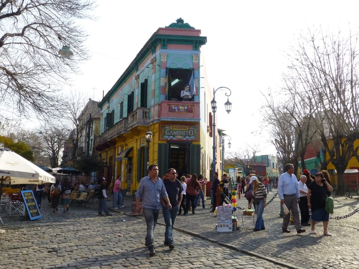 A street corner in the colourful La Boca neighbourhood in Buenos Aires with cobblestone roads and people walking. The corner building is brightly coloured in green, pink, and yellow, with balconies and a mural. The area has outdoor seating, street lamps, and a clear sky.