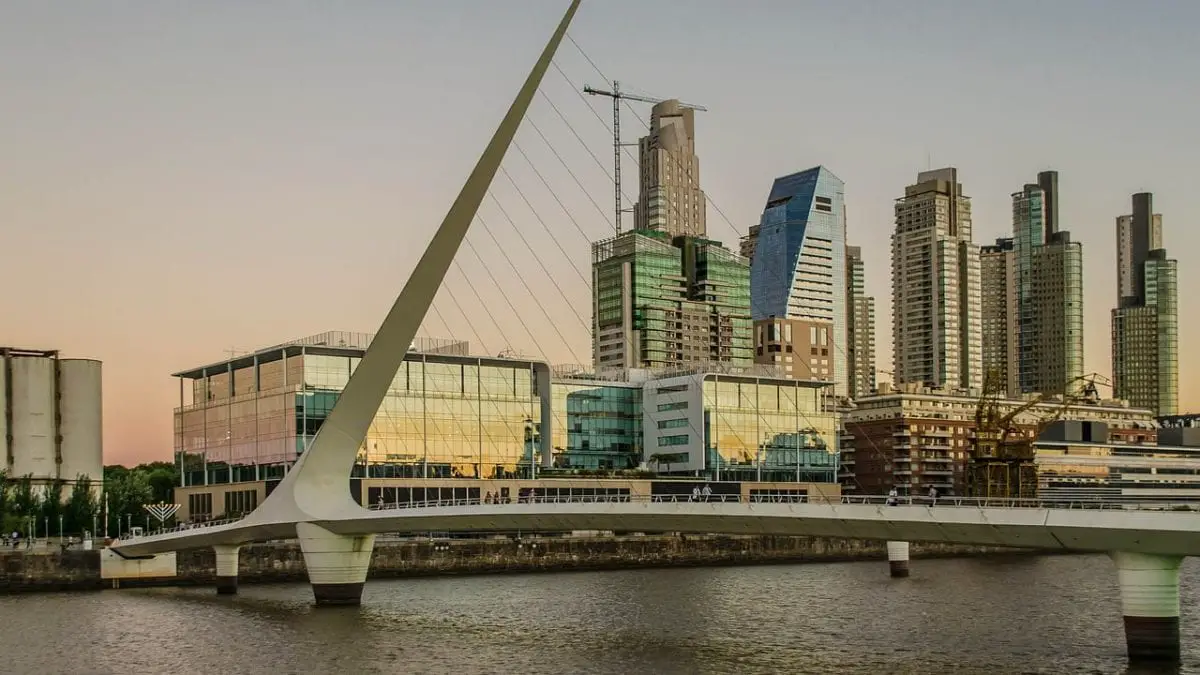 The Puente de Mujeres (Women's Bridge) in Puerto Madero, Buenos Aires. A modern cityscape at dusk with a white, sleekly designed pedestrian bridge in the foreground, crossing a river. Tall buildings with illuminated windows form the skyline, and construction cranes suggest ongoing development. The sky is tinged with the warm hues of sunset.