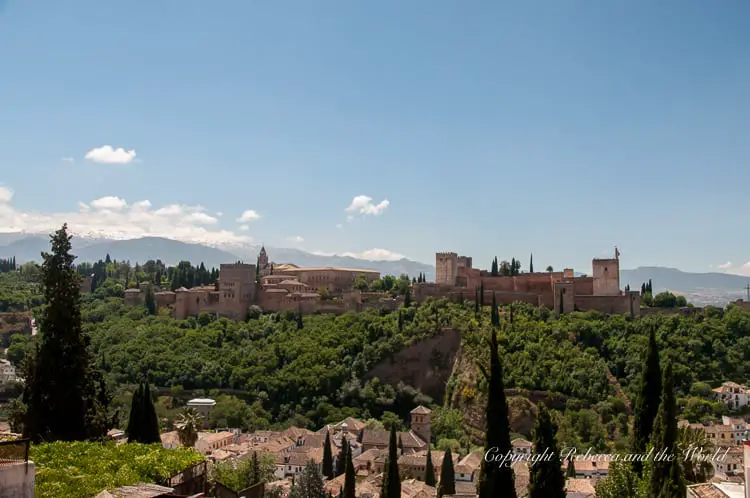 A panoramic view of a historic fortress complex on a hill, surrounded by green trees under a clear blue sky. This is the incredible Alhambra in Granada, Spain, and a highlight of any Southern Spain itinerary.