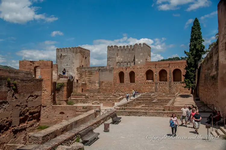 The expansive ruins of the Alhambra's fortifications, featuring large open courtyards with tourists walking around and lush trees dotting the landscape. The Alcazaba is the oldest part of the Alhambra in Granada, Spain. The former forts are still in good condition.