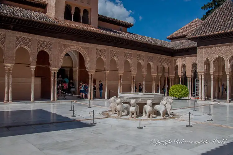 The Patio of the Lions in the Alhambra, showcasing a white marble fountain supported by statues of lions, surrounded by detailed Islamic architecture. The Nasrid Palaces are a must-see when you visit the Alhambra in Granada, Spain.