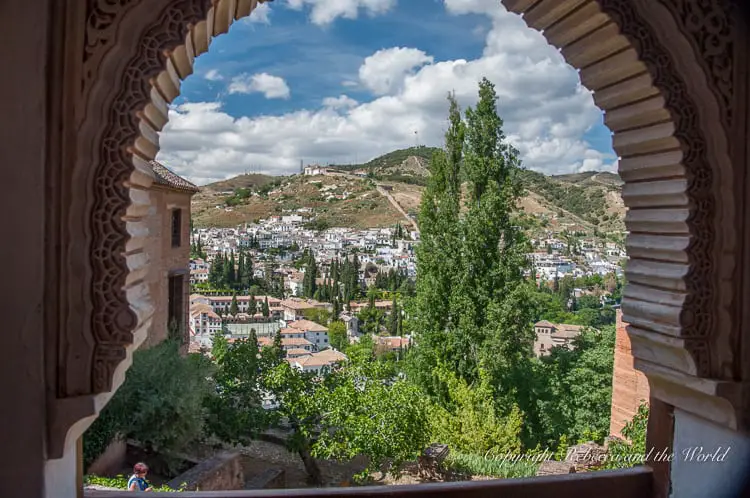 A picturesque view from the Alhambra, through an ornate window arch, overlooking the white buildings of Granada amidst green foliage and hills. There are so many lovely views from the walls and windows of the Alhambra in Granada, Spain.