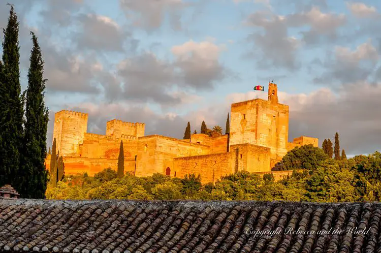 The Alhambra’s robust stone fortress bathed in golden sunlight, with a fluttering Spanish flag atop a tower, and a foreground of dark green cypress trees framing the historic site.