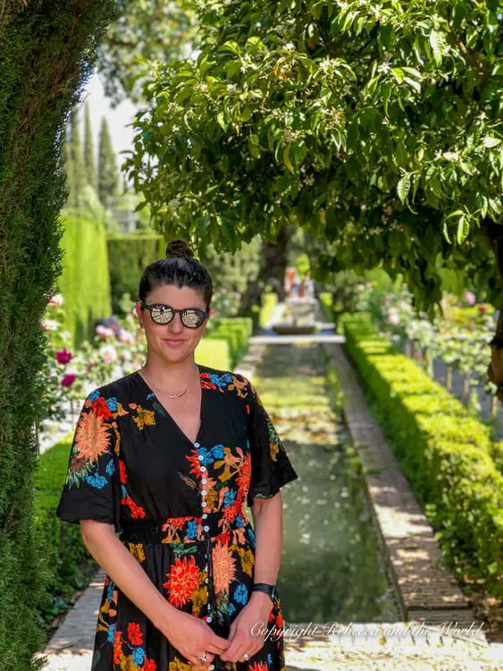 A woman in a floral dress - the author of this article - standing in front of a manicured hedge and a long rectangular reflecting pool in the Generalife gardens, with flowers and trees in the background. Plan to spend at least three hours at the Alhambra. Make sure to take a break in the gorgeous gardens of the Generalife.