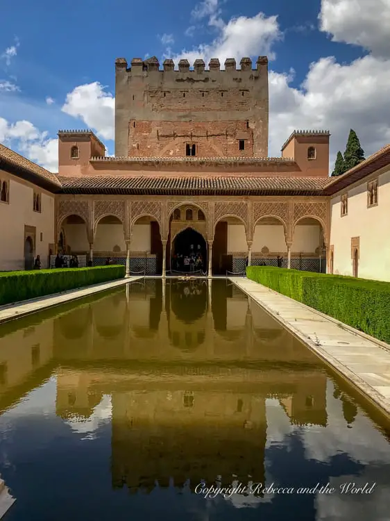 A courtyard with a rectangular reflecting pool, surrounded by ornate arches and a two-storied building with a tower. This is inside the amazing Alhambra complex in Granada, Spain.