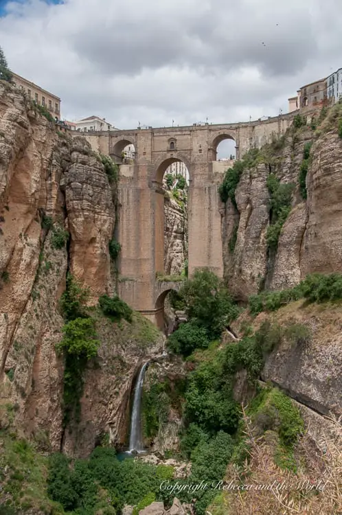 A towering stone bridge with multiple arches spans a deep gorge with a waterfall visible through one of the lower arches. This is the incredible Puente Nuevo in Ronda, Spain.