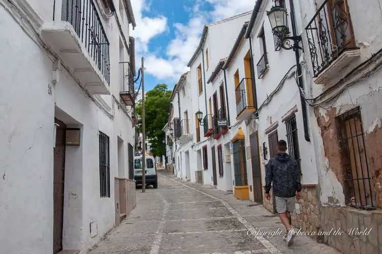 A narrow street in a historic village with traditional white houses and a cobblestone road. A person walks away from the camera.