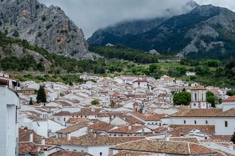 An aerial view of a town with white buildings and red-tiled roofs, nestled in a valley with mountains in the background and overcast skies above. This is one of the pueblos blancos in Andalucia, Spain.