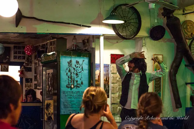 A dimly lit bar with a performer in traditional flamenco attire, surrounded by an audience and posters on the walls.