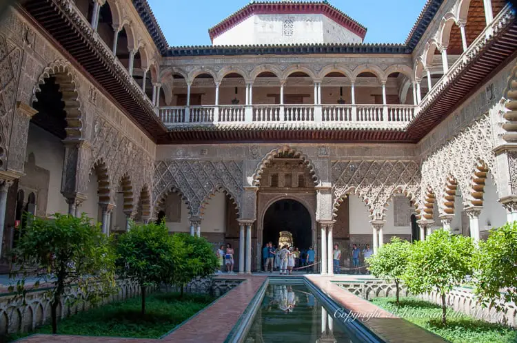 A courtyard with a long reflective pool, surrounded by intricately carved archways and a multi-level building with visitors walking around. This is the inside of the Real Alcazar in Seville, Spain.