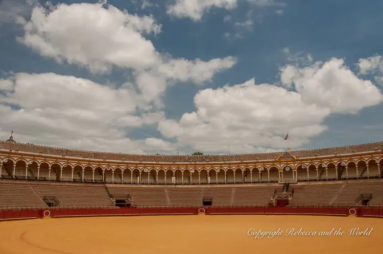 An empty bullring with sandy ground and tiers of seating under a partly cloudy sky.