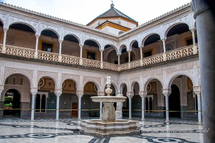 A serene courtyard featuring a marble fountain in the center, surrounded by arched walkways and ornate tiled walls. This is the Casa de Pilatos in Seville, Spain.