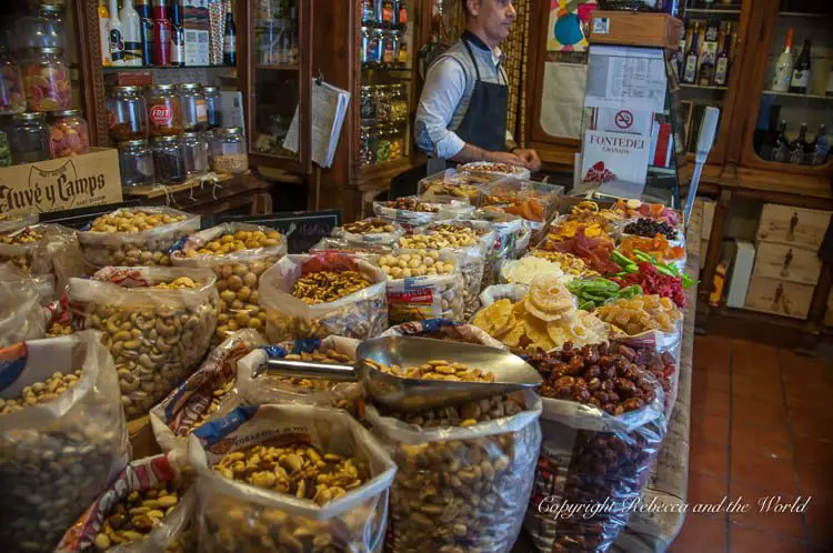 An indoor market stall with an array of nuts, dried fruits, and snacks displayed in open bags. A person behind the counter appears to be working. This is the oldest store in Spain!