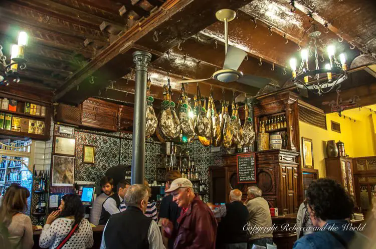 A traditional tavern with hanging hams, wooden bar counters, and patrons engaging in conversation. This is the interior of El Rinconcillo, the oldest bar in Seville, Spain.