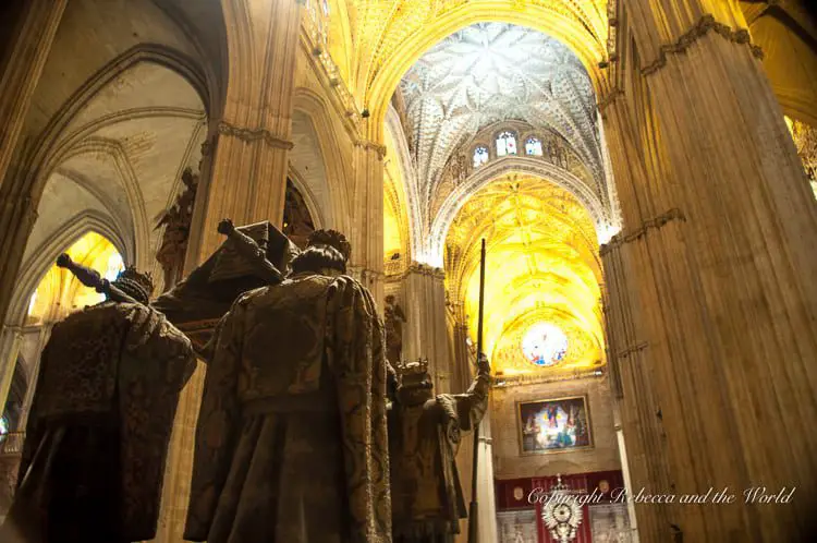 Interior of a cathedral with tall, ornate golden ceilings and statues of figures in medieval attire, with a glowing altar in the background. This is the Seville Cathedral in Andalucia, Spain.