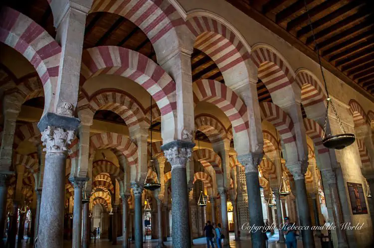 The interior of a mosque with red and white striped arches, columns, and hanging lamps, with visitors in the background. This is the interior of La Mezquita in Cordoba, Spain.