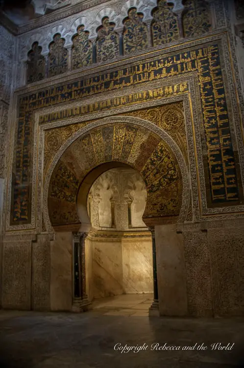 An ornate archway with intricate golden and blue patterns on the walls, leading into a dimly lit alcove. This is the interior of La Mezquita in Cordoba, Spain.