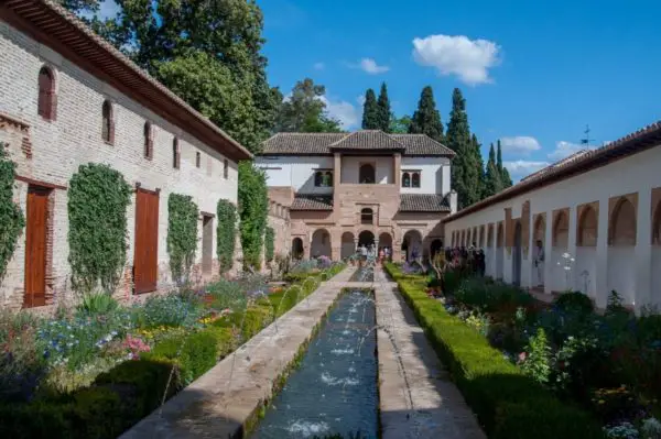 The Generalife's tranquil garden pathway flanked by vibrant flower beds, hedgerows, and a long central water channel, leading to an ornate pavilion. The Generalife in the Alhambra, Spain, is filled with gorgeous gardens.
