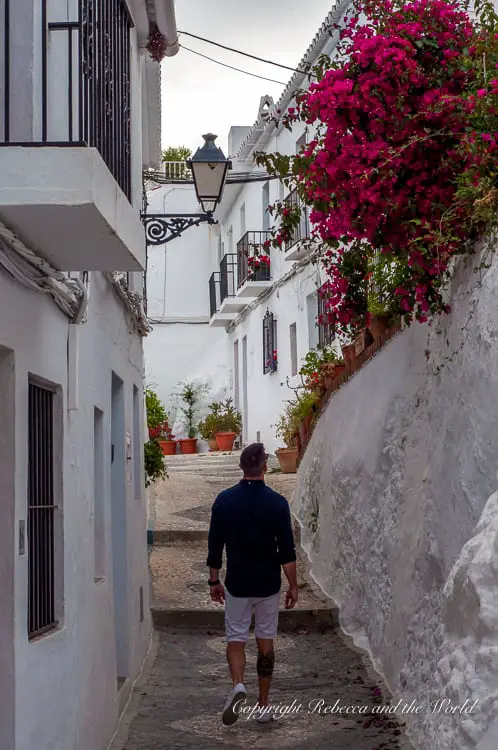 A person walks down a narrow, whitewashed alley with a lush pink bougainvillea hanging over a wall on the right side. This is one of the 