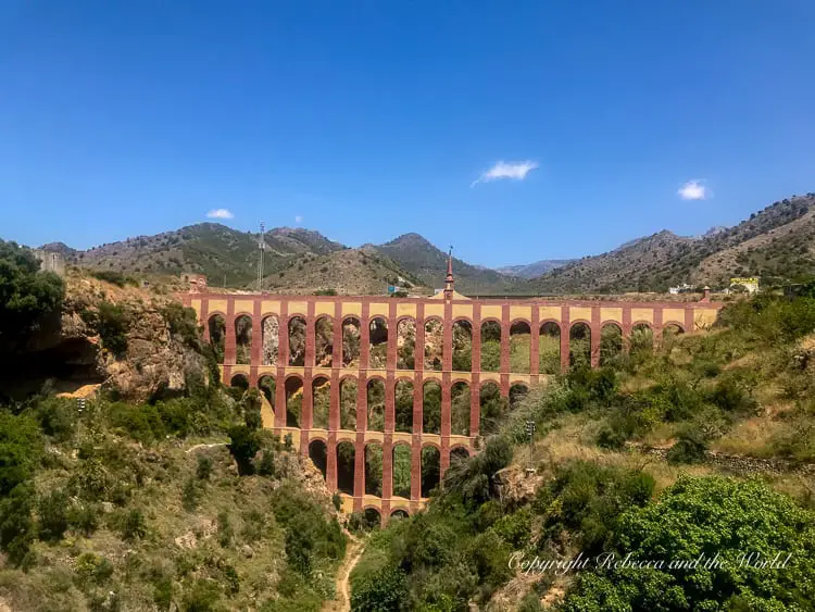 A large, multi-arched aqueduct bridge spans a valley, set against a backdrop of hills and clear blue sky. This aqueduct is just outside of Nerja in Andalucia, Spain.