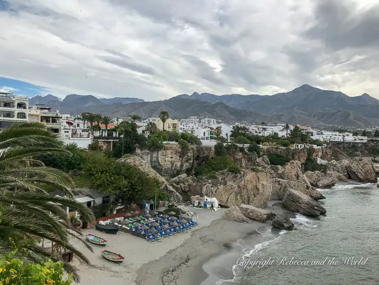 A coastal view of a beach with umbrellas and loungers, with white buildings and mountains in the background. This is Nerja, one of the towns on this recommended Andalucia road trip itinerary.